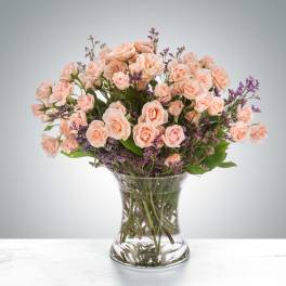 Peach spray roses with purple filler flowers arranged in a clear glass vase.