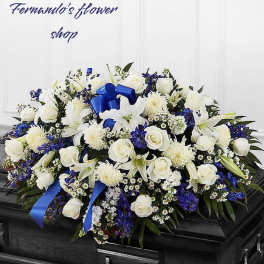 White and blue funeral casket spray with roses, lilies, and ribbon on a dark wood casket