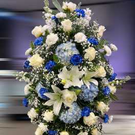 Tall standing spray of white lilies, roses, carnations and blue hydrangeas with bright blue accent blooms.