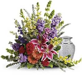 Sympathy floral spray of lilies, roses, and mixed flowers arranged beside a decorative cremation urn.