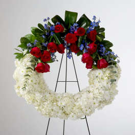 Standing wreath of white flowers with a top spray of red roses and blue blooms on a metal easel.
