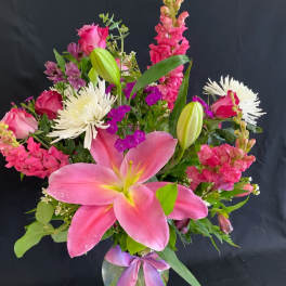 Tall mixed arrangement of pink lilies, roses, snapdragons and white mums in a clear vase with a ribbon bow