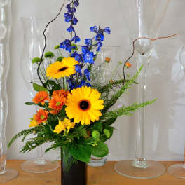 Tall arrangement of yellow gerbera, orange mums, and blue delphinium in a black rectangular vase