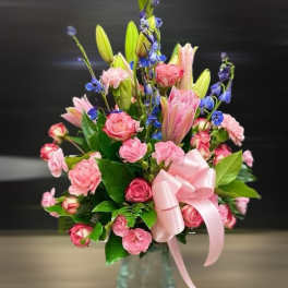 Pink roses and lilies with blue accent flowers in a glass vase with a pink bow