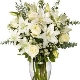 White lilies, roses, and small white blossoms arranged in a clear glass vase