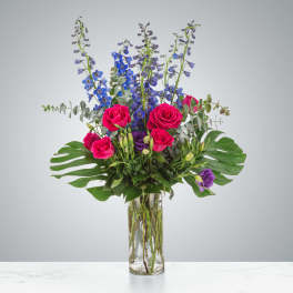 Tall arrangement of hot pink roses, blue delphinium, and purple blooms in a clear glass cylinder vase