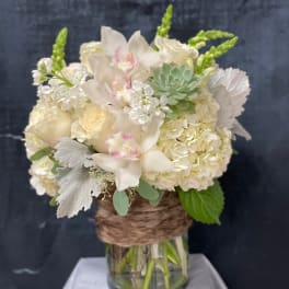 White hydrangeas, roses, orchids, and a succulent in a glass vase wrapped with brown twine