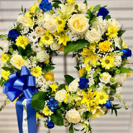 Standing wreath of white, yellow, and blue flowers with a large blue bow on an easel.