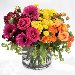 Bright arrangement of pink gerbera daisies, roses, and yellow and orange flowers in a clear glass vase