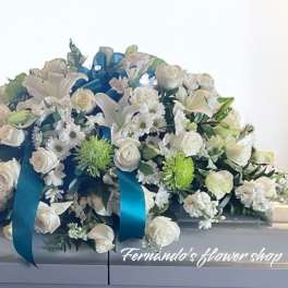 White and green casket spray of roses, lilies, and mums with teal ribbon on a white casket.