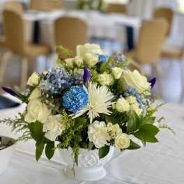 Low centerpiece of white roses, carnations, and blue accents in a white pedestal bowl