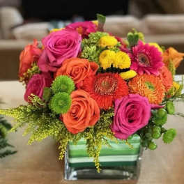 Bright arrangement of pink roses, orange blooms, and yellow flowers in a square glass vase