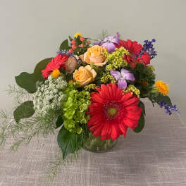 Mixed bouquet with red gerbera daisies, peach roses, and purple flowers in a glass vase