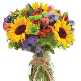 Bright mixed bouquet of sunflowers, tulips, and purple daisies in a glass vase