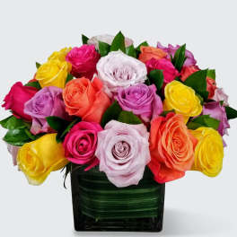Compact arrangement of multicolored roses in a leaf-lined square glass vase