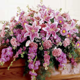 Pink and lavender casket spray with lilies, carnations, and daisies on a wooden casket.