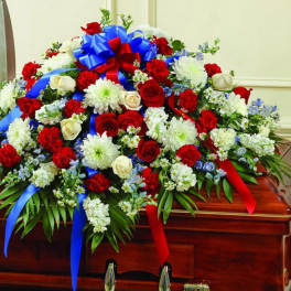 Patriotic red, white, and blue casket spray with roses, carnations, mums and ribbons on a wooden casket
