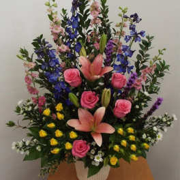 Tall mixed arrangement with pink lilies, pink and yellow roses, and blue delphinium in a white container