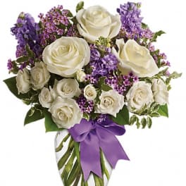 White roses and purple flowers arranged in a glass vase with a purple ribbon