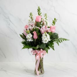 Pink roses and white flowers arranged in a glass vase with a pink ribbon