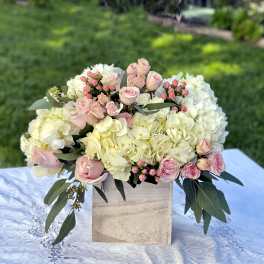 Low arrangement of cream hydrangeas and pale pink roses in a wooden cube vase on a lace-covered table.