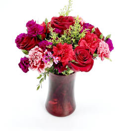 Red and pink roses and carnations arranged with purple blooms in a red glass vase