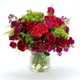 Vase arrangement of red roses, pink carnations, and purple blooms with bright green accents in a clear glass cylinder.