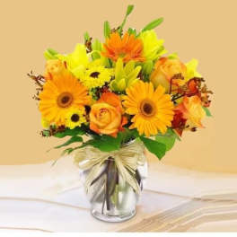 Bright orange gerberas, yellow lilies, and roses in a clear glass vase with a raffia bow