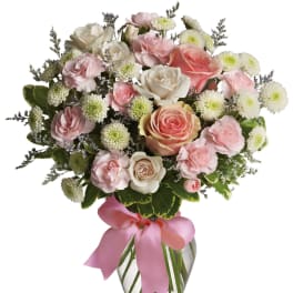 Pink and white roses, carnations, and white mums in a clear glass vase with a pink ribbon bow