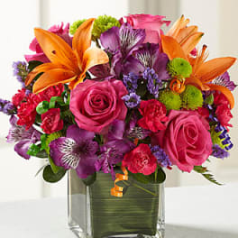 Pink and purple bouquet with orange lilies in a square glass vase
