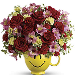 Red roses and pink flowers arranged in a yellow smiley-face mug
