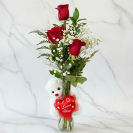 Three red roses in a glass vase with a teddy bear and heart tag