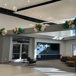 Balloon clusters in green, white, and gold hang from a lobby beam.