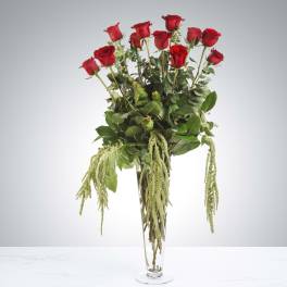 Tall arrangement of long-stem red roses with trailing green accents in a clear glass vase