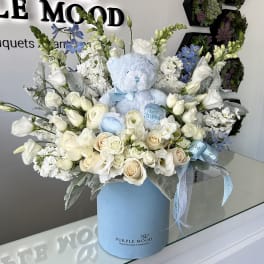 Blue teddy bear in a white and blue flower arrangement in a round box