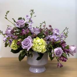 Lavender rose arrangement in a white pedestal vase with purple blooms