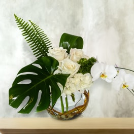 White roses and orchids arranged in a glass bowl with large tropical leaves