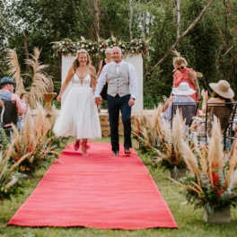 Outdoor wedding aisle with pampas grass arrangements and a flower-topped arch behind a couple on a red carpet.