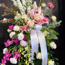 Tall standing spray of pink lilies, roses, and white flowers with a white ribbon on an easel.