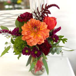 Compact bouquet with orange dahlia and rose, burgundy blooms, and green accents in a clear glass vase.