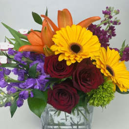 Bouquet of yellow gerbera daisies, red roses, orange lilies, and purple flowers in a glass vase
