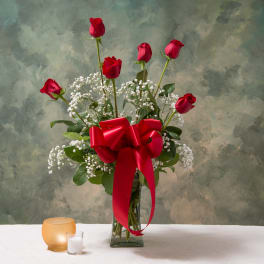 Tall arrangement of red roses and white filler flowers in a glass vase with a large red bow.