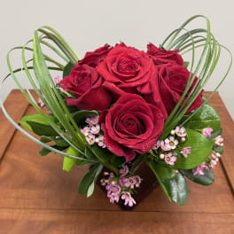 Compact arrangement of red roses with pink filler flowers in a red cube vase framed by looping grasses.