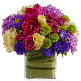 Colorful arrangement of pink, purple, yellow, and green flowers in a clear square glass vase