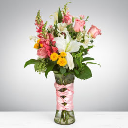 Tall mixed bouquet of pink roses, white lilies, yellow spray roses and snapdragons in a glass vase wrapped with pink ribbon