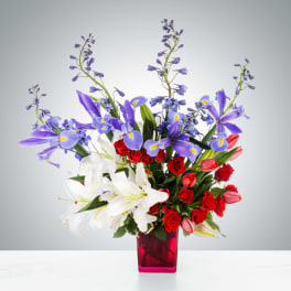 Tall arrangement of purple irises, white lilies, red roses, and tulips in a bright pink glass vase