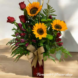 Arrangement of yellow sunflowers, red roses, and purple blooms in a red vase with a burlap bow
