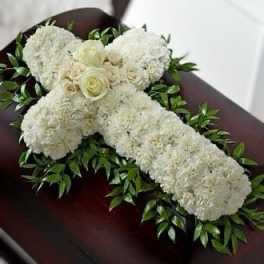 White floral cross with roses and carnations on greenery displayed on a dark wood casket