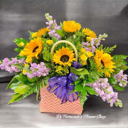 Yellow sunflowers, yellow spray roses, and purple flowers arranged in a pink patterned tote with a purple bow.