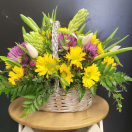 Basket arrangement of yellow daisies, purple blooms, and pale tulips